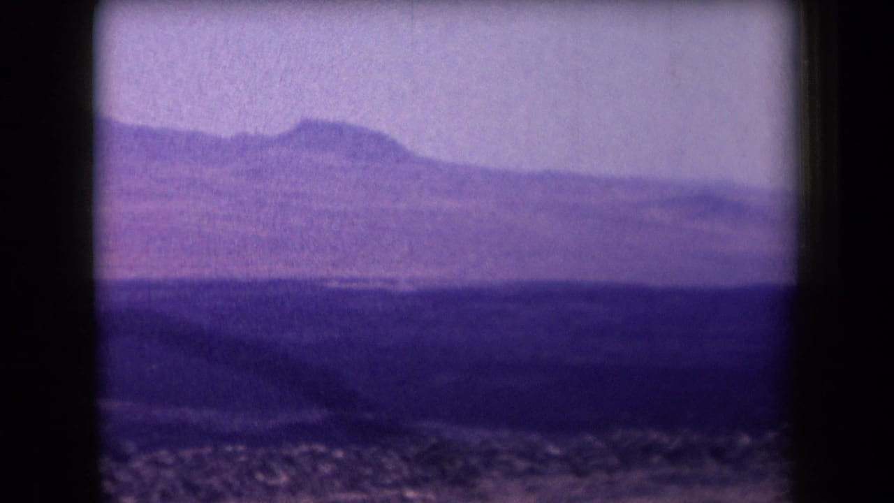 1958 Arizona home-movie frame showing a purple-tinted desert plain, rocky foreground, and distant mountain ridge beneath a hazy sky.