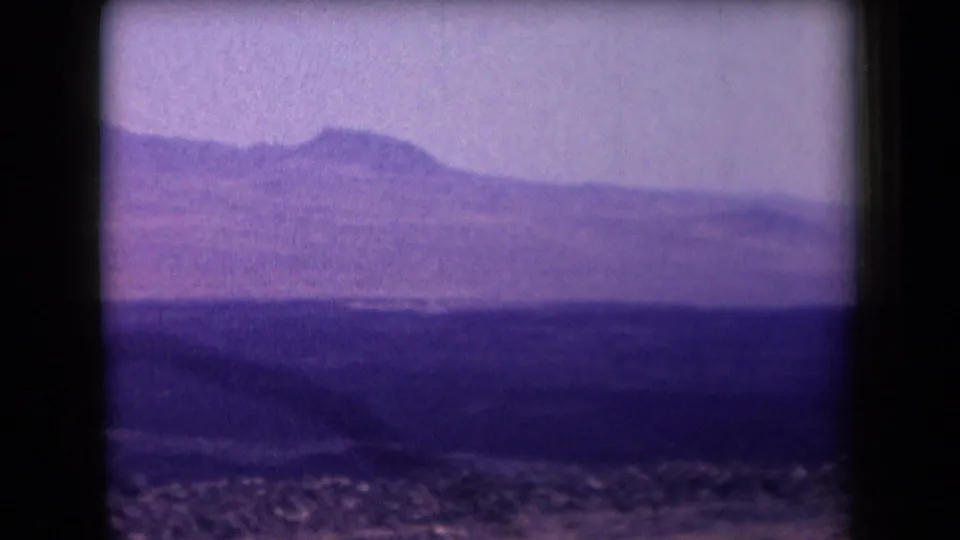 1958 Arizona home-movie frame showing a purple-tinted desert plain, rocky foreground, and distant mountain ridge beneath a hazy sky.