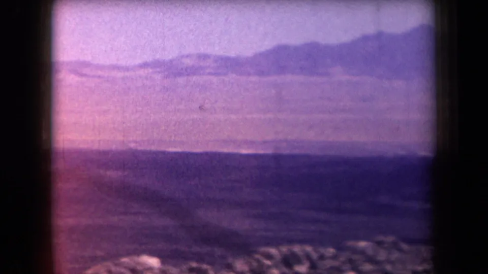 1958 Arizona home-movie still showing layered desert terrain, low mountains, rocky foreground, and a pink-purple color cast.
