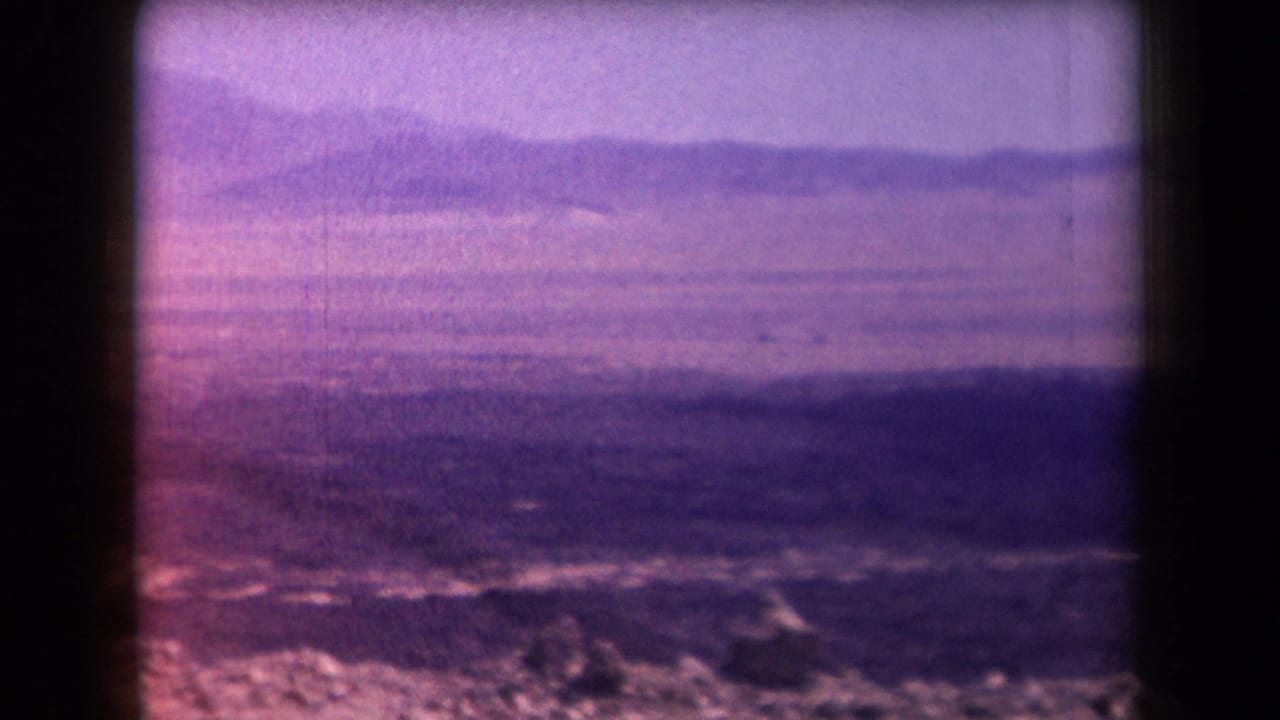 1958 Arizona 8mm frame showing a hazy mountain landscape through dark window bars, with purple color shift and dry foreground texture.