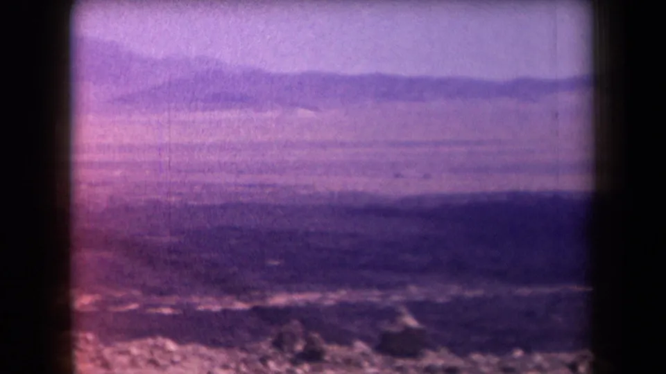 1958 Arizona 8mm frame showing a hazy mountain landscape through dark window bars, with purple color shift and dry foreground texture.