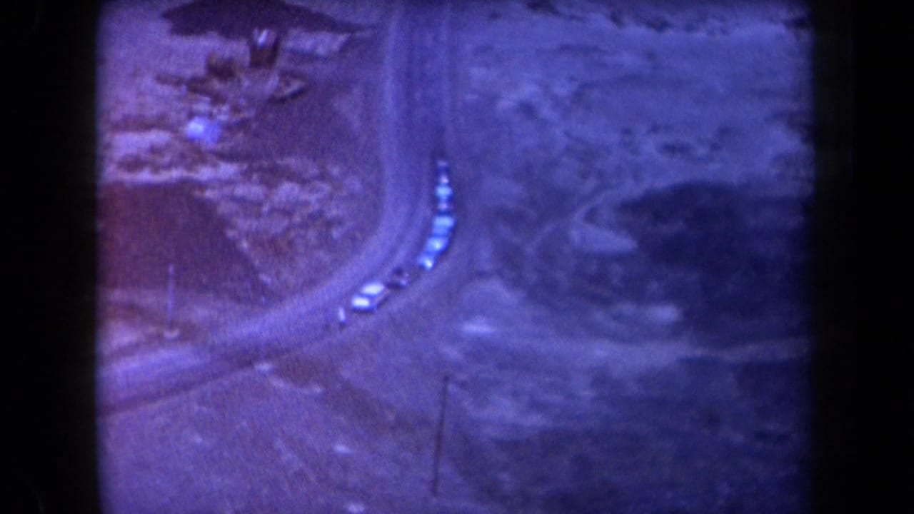 1958 Arizona home-movie frame showing several cars rounding a curved dirt road in rugged desert terrain with a blue-purple cast.