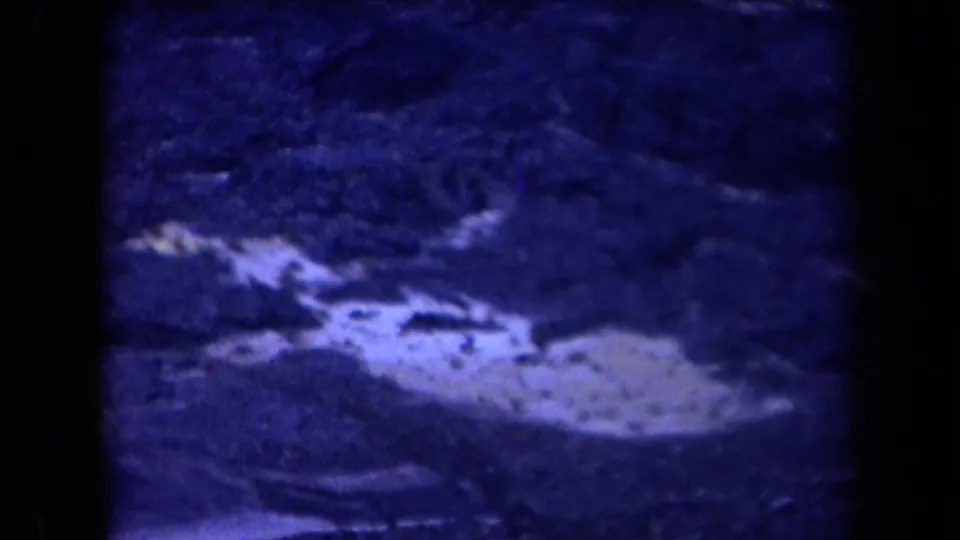 Blue-tinted 8mm frame of rocky Arizona desert terrain below the road, with pale ground and scrub visible in soft focus.