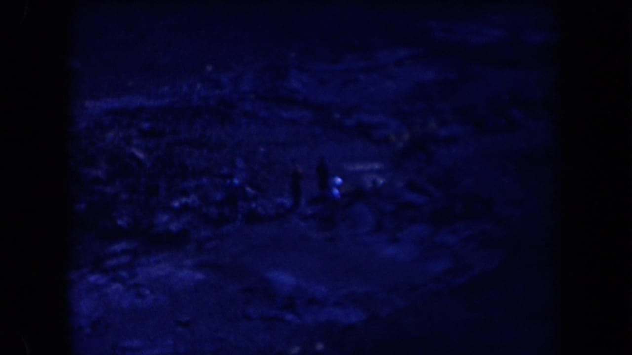 Blue-tinted 1958 Arizona 8mm frame showing a dim open landscape with small human figures and heavy film borders.