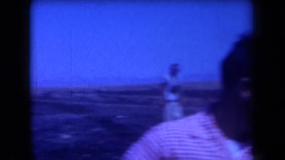 Blurred close view of the checkered-shirt man in blue-tinted 1958 Arizona 8mm footage, with a distant figure behind him.