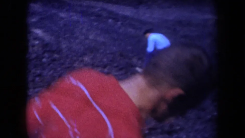 A person in a red shirt with light striping bends forward on a dark coal slope in blue-shifted 8mm footage.