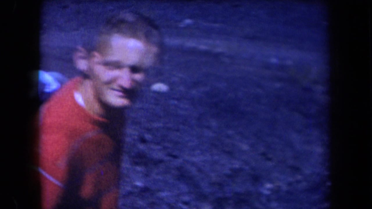 In 1958 Arizona 8mm footage, an adult man in a red shirt looks up from rocky ground toward the high-angle camera.