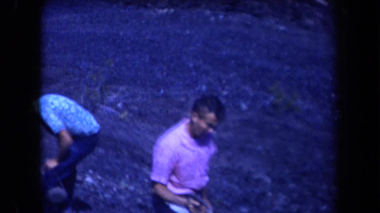 Two adult men stand or bend on rocky ground, one wearing a pink shirt.
