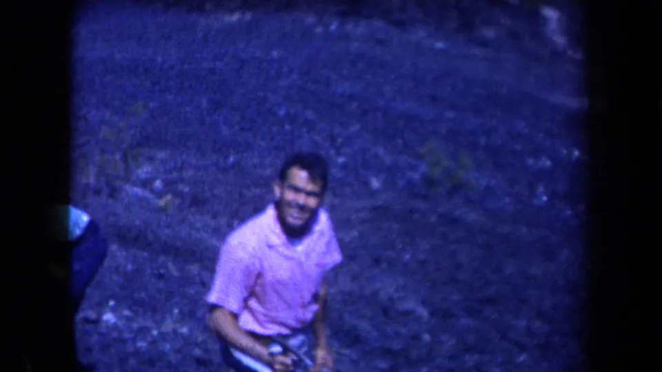 A man in a pink polo shirt smiles up at the handheld 8mm camera in a rocky Arizona outdoor setting.