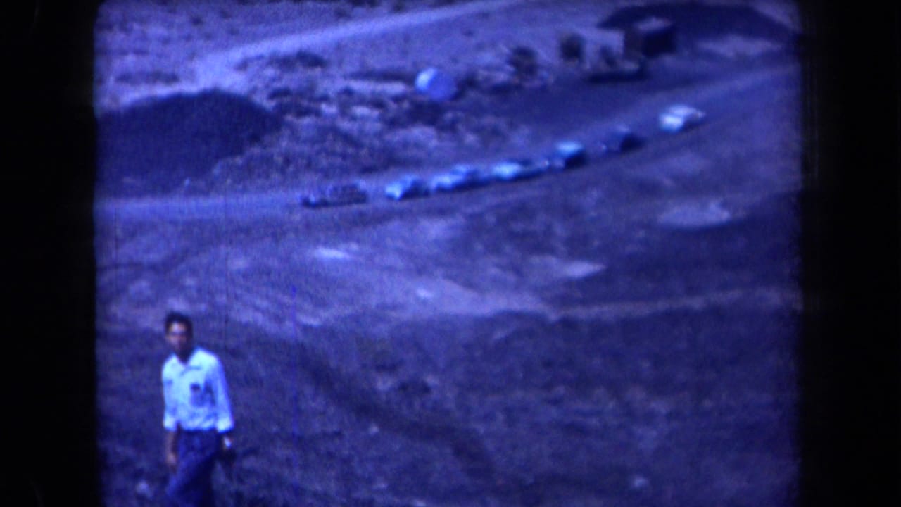 1958 Arizona 8mm frame showing a young man near the camera and several cars on a winding dirt road across arid hills.