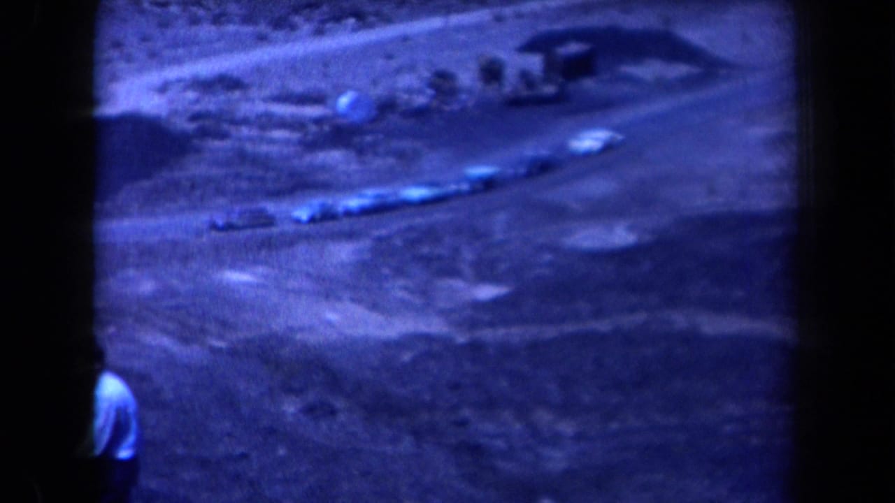 Cars follow a curving dirt road through hills as a person appears near the lower edge.