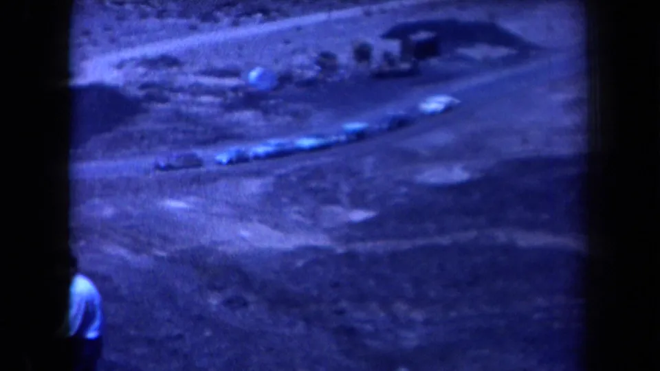 Blue-tinted 1958 Arizona home-movie frame with cars on a hillside dirt road and a person partly visible near the foreground.
