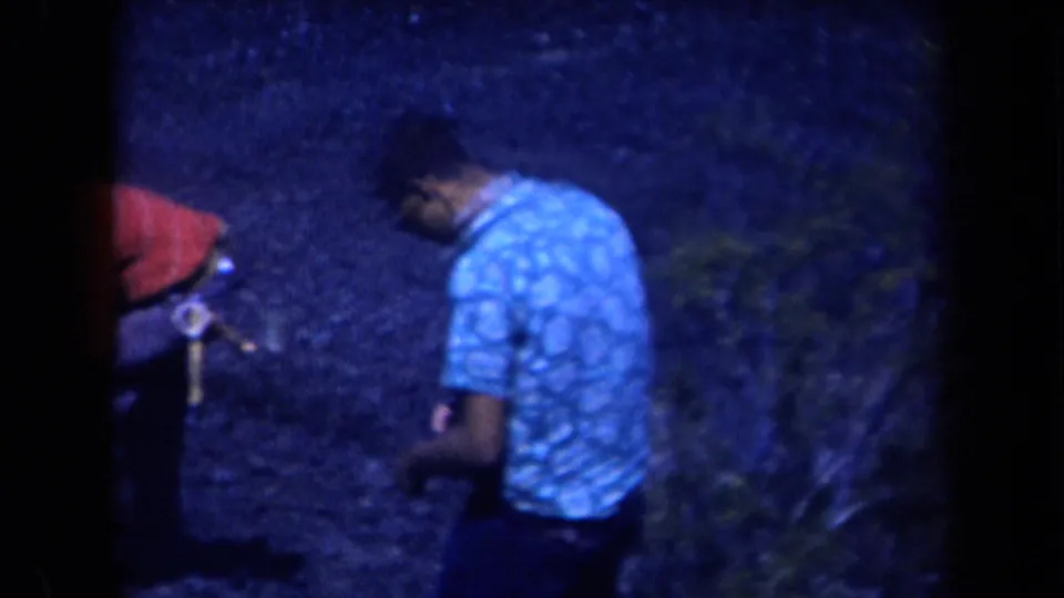 A man in a plaid short-sleeved shirt looks down near rocky ground, with part of a red object visible at frame edge.