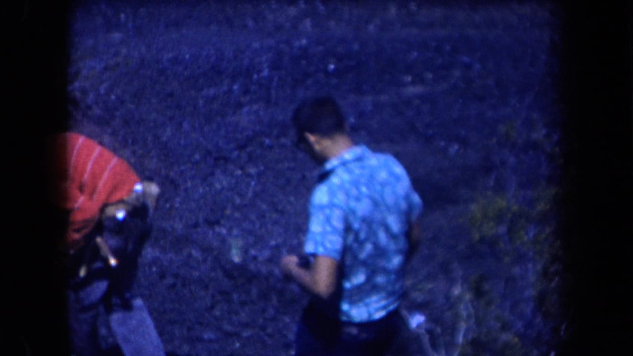 The two adult men continue across rocky Arizona terrain, partly turned away from the camera in blue-tinted 8mm footage.