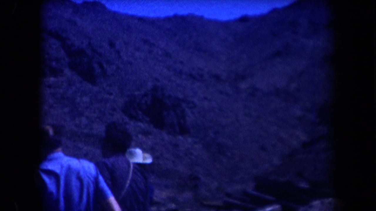 1958 Arizona 8mm frame showing adults near a car at the base of a rugged mountain slope, with dark blue low-light exposure.