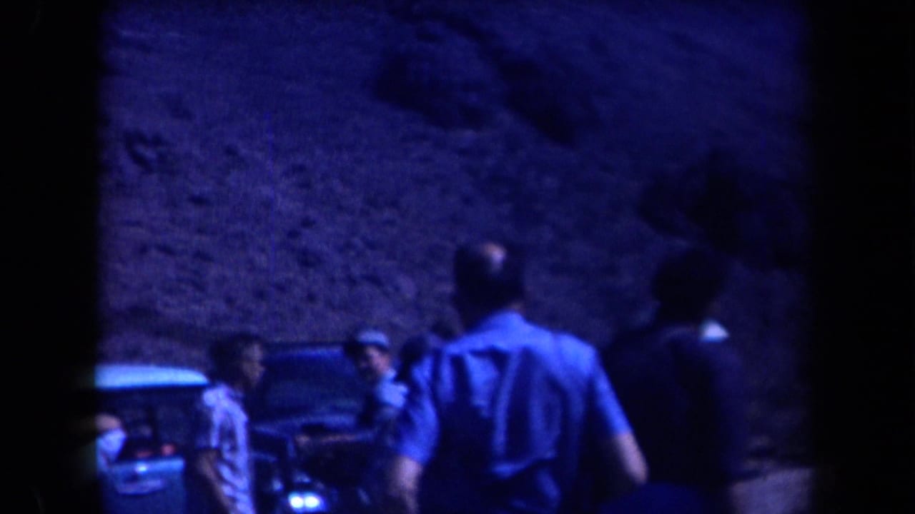 Men stand near a car and a ladder at the base of a rocky mountain slope.