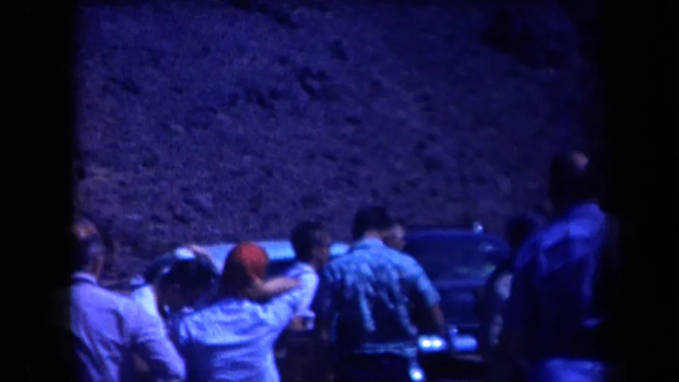 1958 Arizona 8mm frame showing several adults, including a person in a red head covering, gathered near a car in a rugged valley.