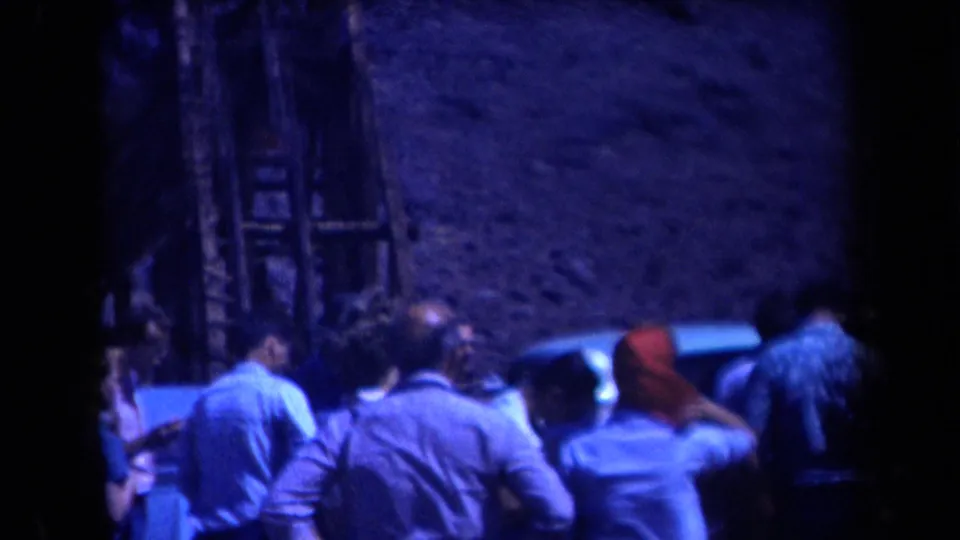 1958 Arizona home-movie frame with adults near a car, a visible ladder, and a rugged mountain backdrop in blue low-light 8mm color.