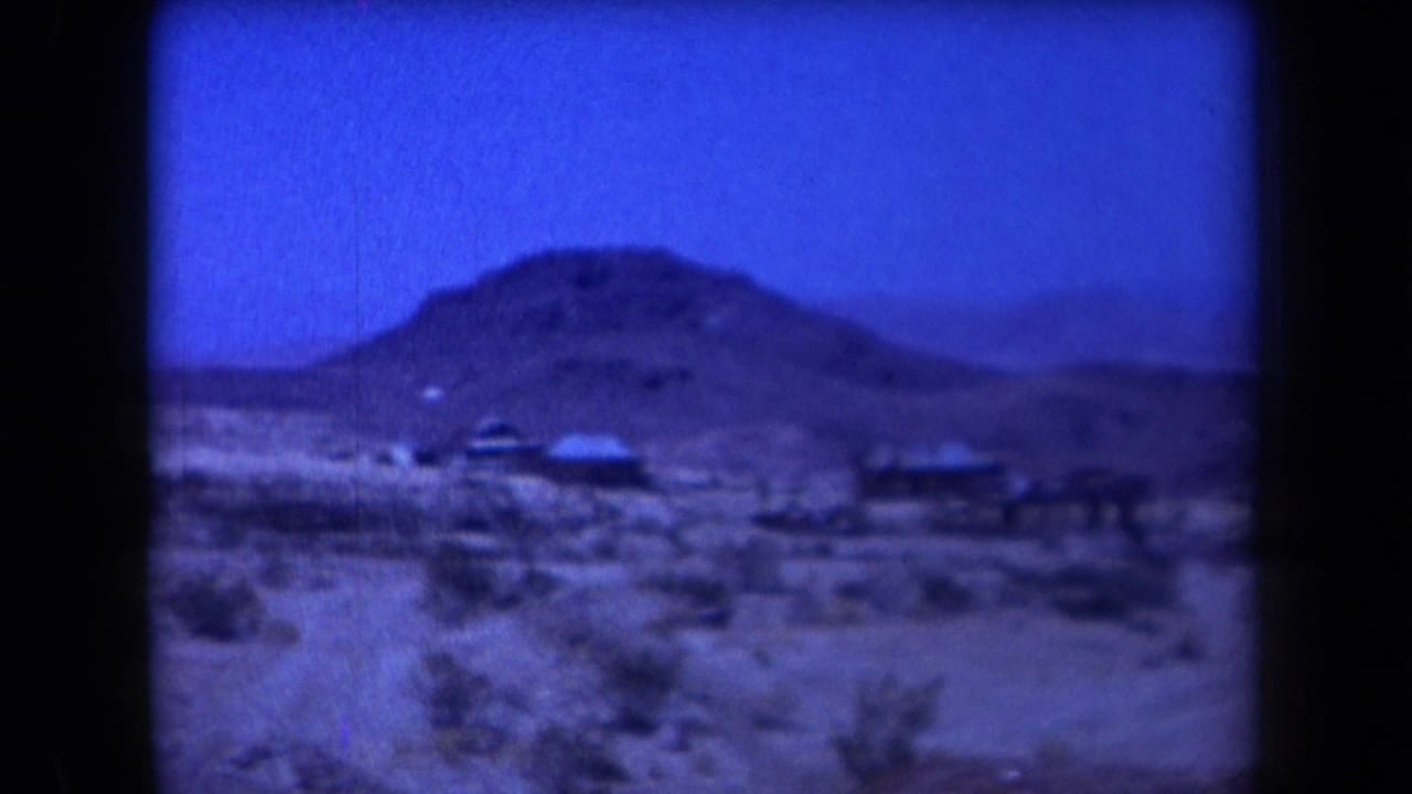 Remote desert view with low hills, scrub, small structures, and dark film edges.