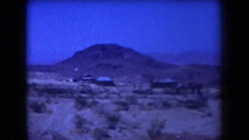 1958 Arizona 8mm frame showing a remote desert landscape with low hills, sparse scrub, and small distant structures.
