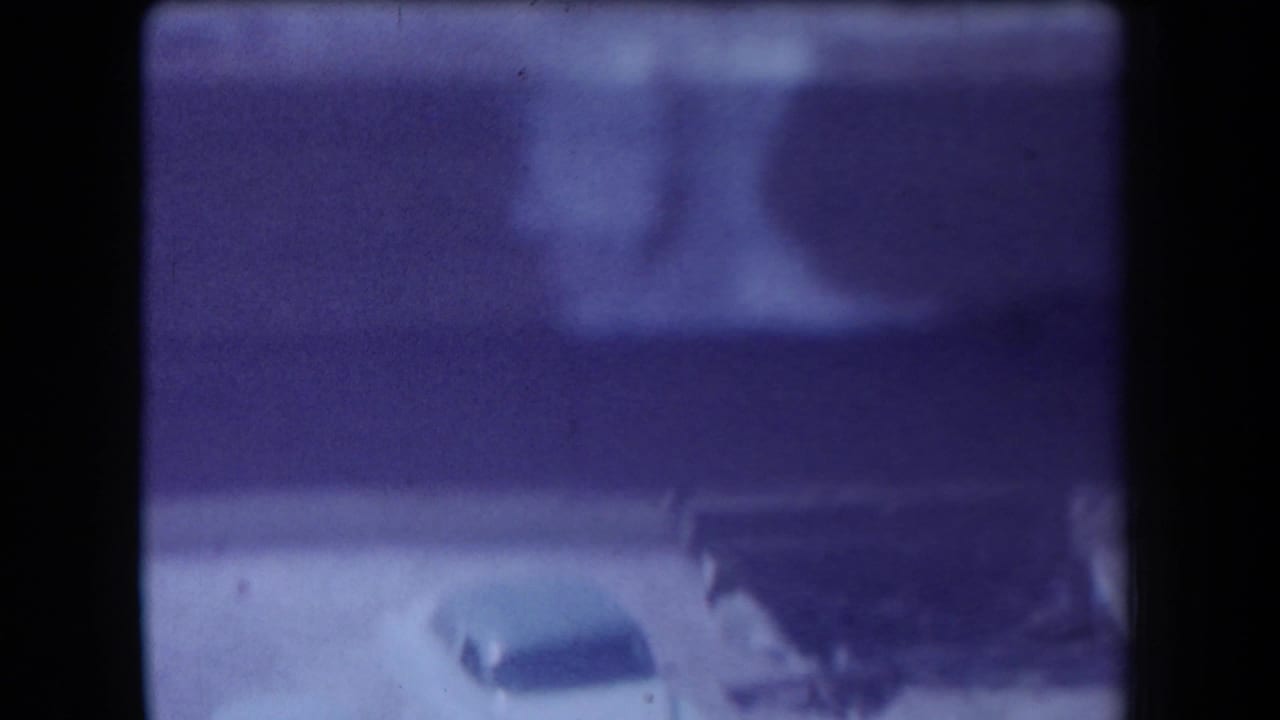 1958 Arizona home-movie frame with two older cars near a low structure or shelter, seen through grain and a purple-blue color cast.