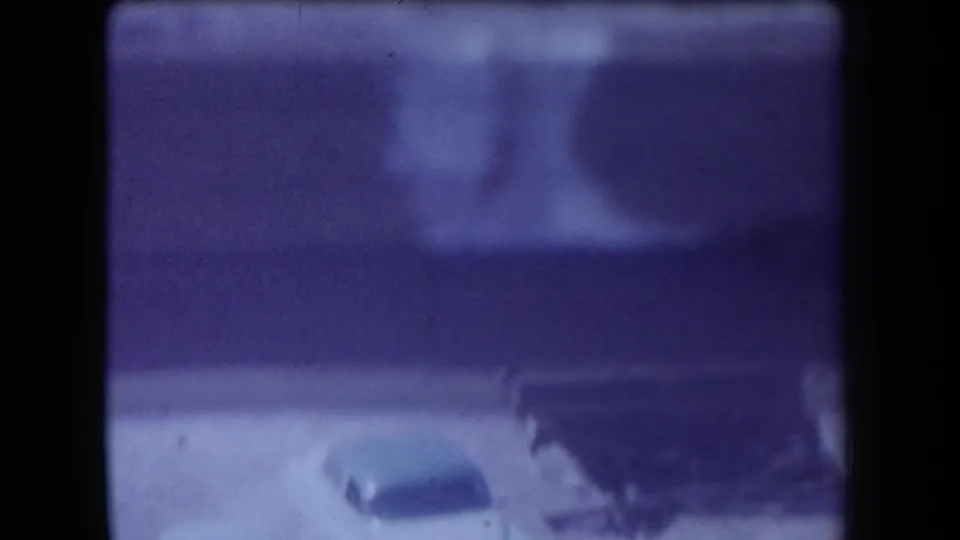 1958 Arizona home-movie frame with two older cars near a low structure or shelter, seen through grain and a purple-blue color cast.