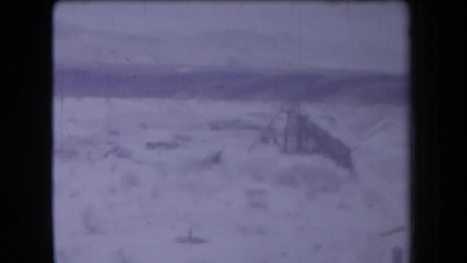 1958 Arizona frame showing an adult figure walking near scrub and a low structure-like form in a purple-tinted desert landscape.