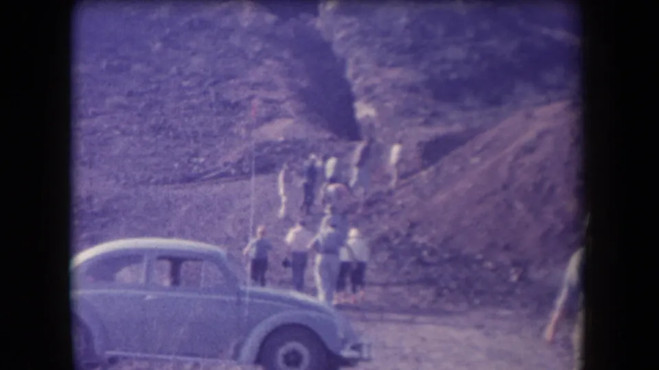 Several people in casual clothing move up a rocky Arizona hillside beside a light blue rounded car in faded 1958 8mm footage.