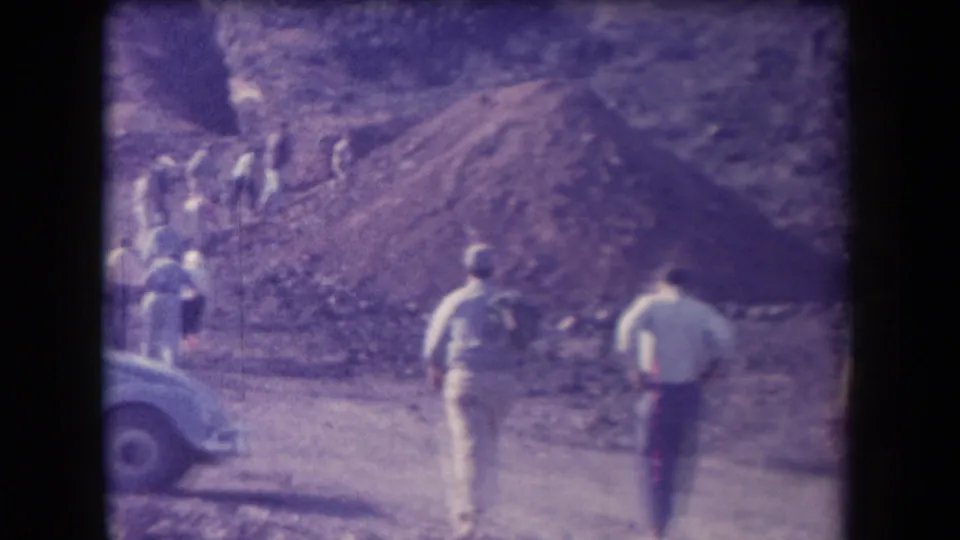 Two figures watch from the lower road while a group climbs the rocky Arizona slope in soft, purple-tinted 1958 home-movie footage.