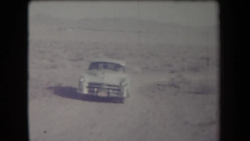 White mid-century car appears on a pale desert road or track in Arizona, with scrubland and low hills in the distance.