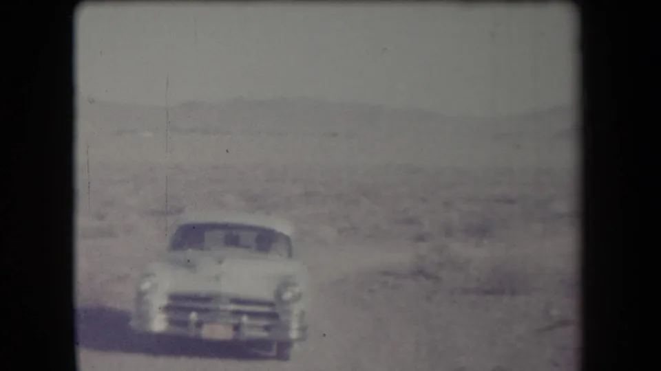 Closer view of a white mid-century car approaching through an arid Arizona landscape, with faded color and visible film wear.