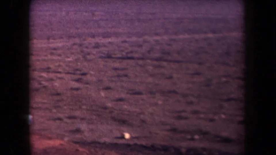 1958 Arizona 8mm frame showing broad desert terrain with a small parked car near the right side and distant haze beyond.