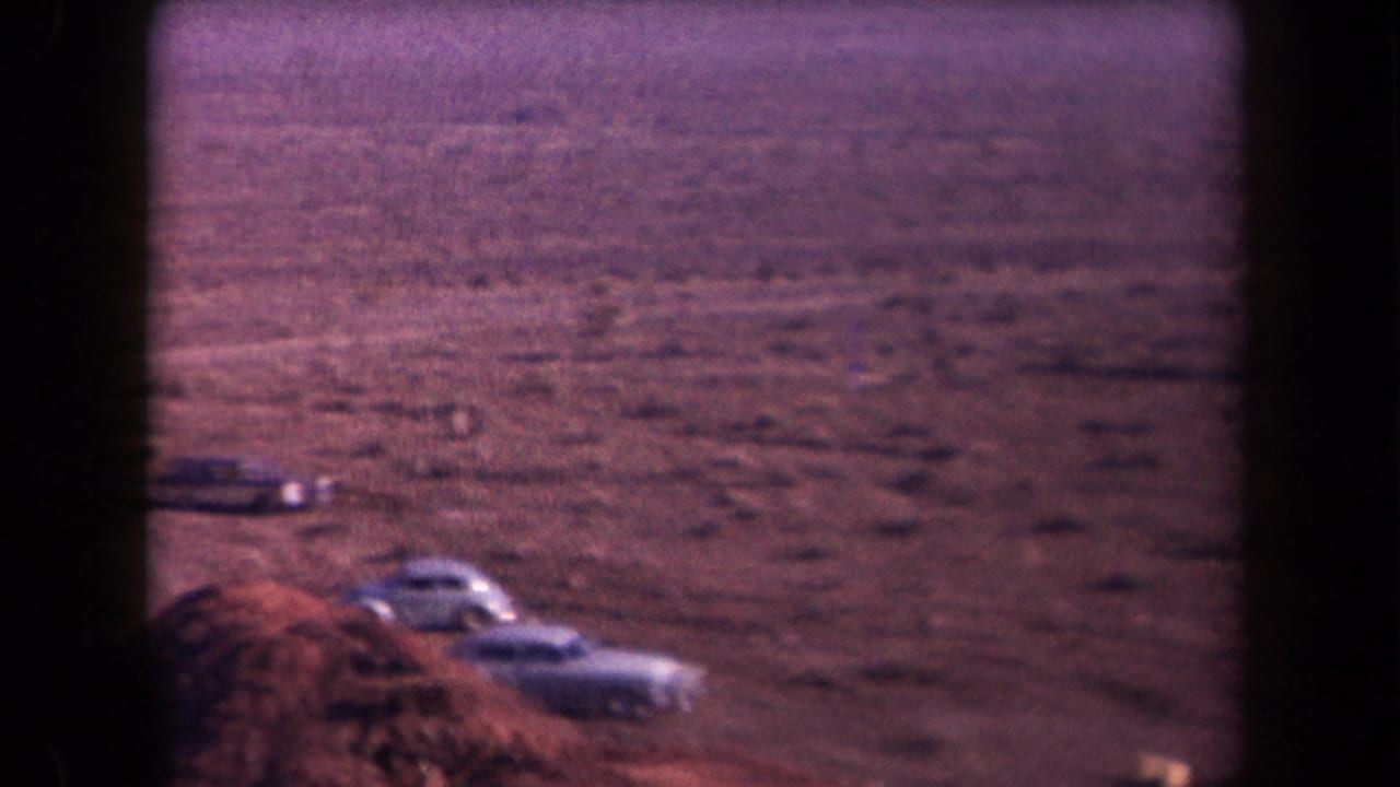 1958 Arizona 8mm frame showing parked cars beside a dirt road in a broad, dry desert landscape.