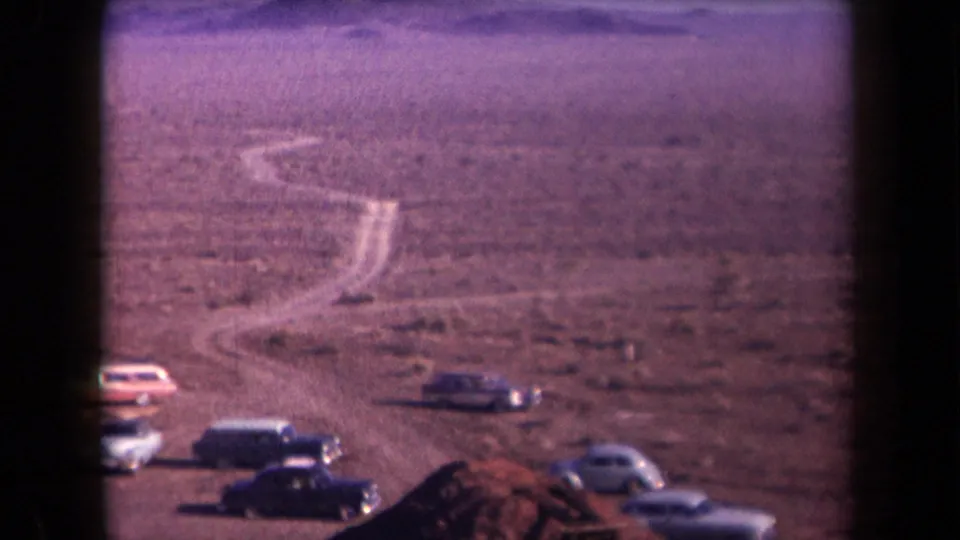 1958 Arizona desert scene with classic cars parked near a winding unpaved road and distant low mountains.