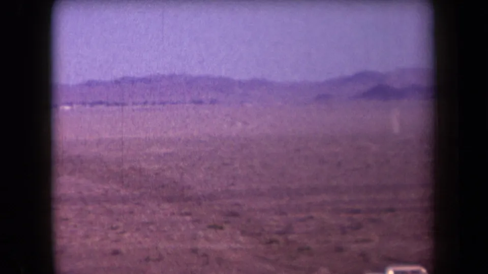 1958 Arizona 8mm frame looking across dry open desert toward distant mountains with a roadside car partly visible.