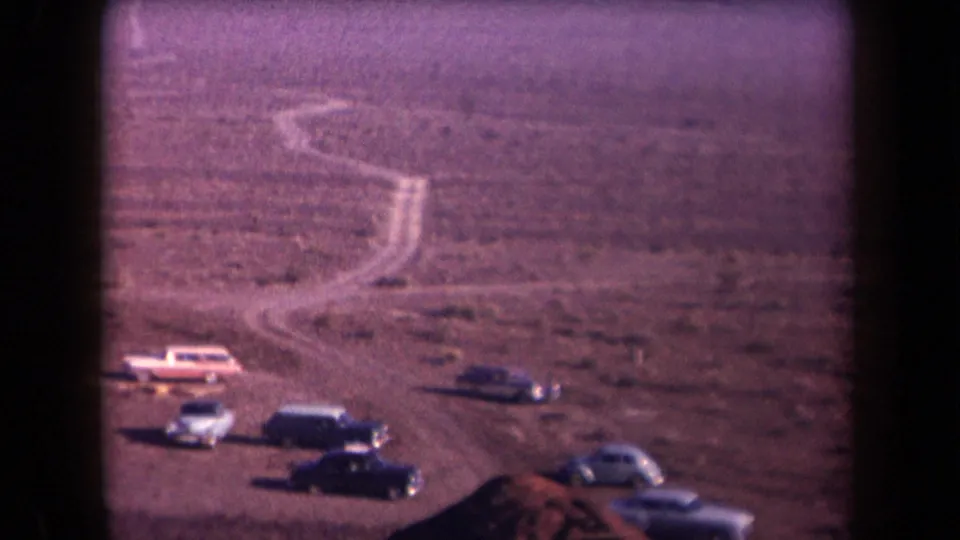 Home-movie frame from 1958 Arizona showing parked mid-century cars and a pale dirt track crossing open desert.