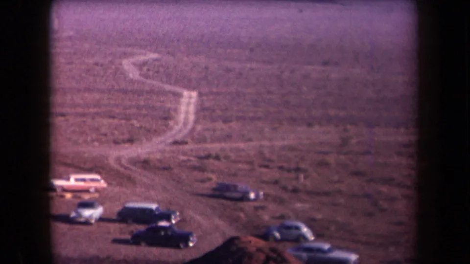 1958 Arizona 8mm frame with several parked cars, a winding desert path, scrubland, and distant hazy mountains.