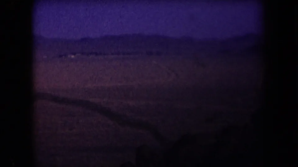 1958 Arizona 8mm frame with open desert, low hills, and a faint distant structure or light patch near the horizon.