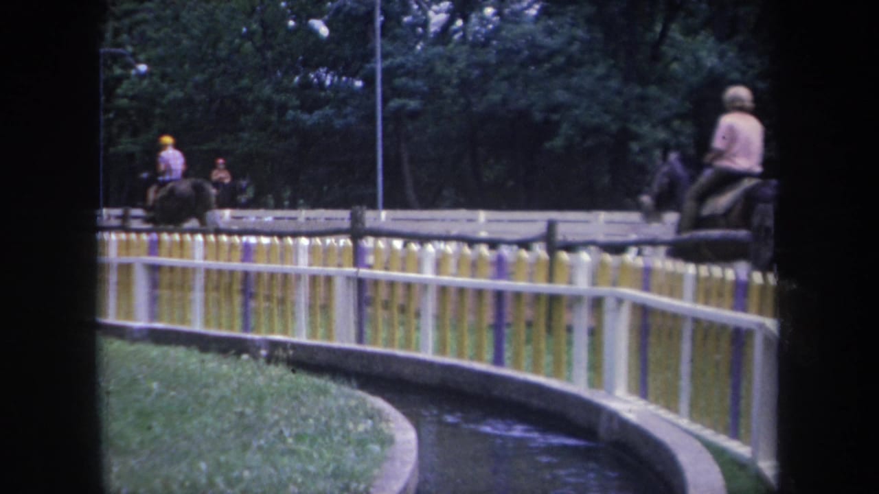 Several riders on horses pass along a fenced track with trees behind them.
