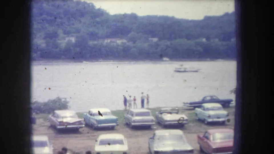 1968 Michigan 8mm frame showing late-1960s cars parked by a river, with people near the shore, wooded hills, and a small boat on the water.