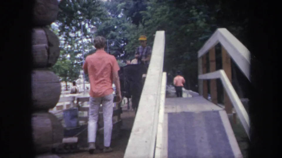 1968 Michigan home-movie frame of a man walking near a wooden bridge, with a horse rider, rail fence, and trees behind.