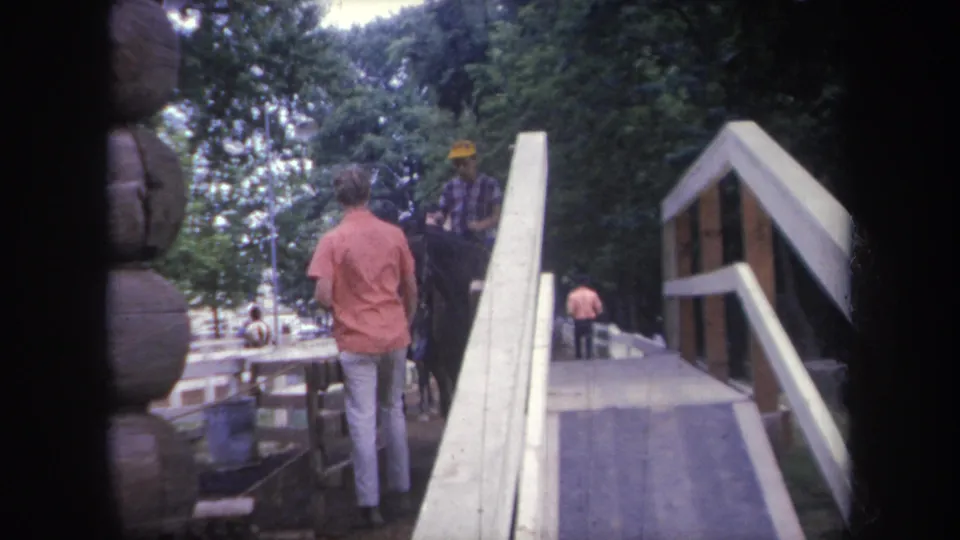 1968 Michigan home-movie frame with a horse and rider beside a ramp, framed by logs, railing, and trees.