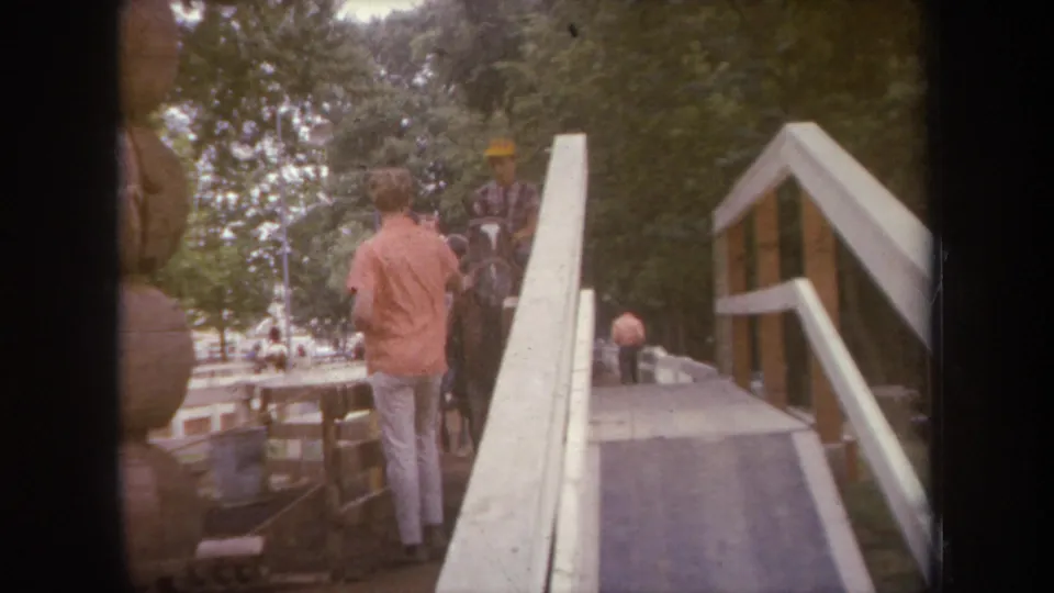 1968 Michigan 8mm frame showing a man, horse, and yellow-hatted rider moving near a wooden ramp with trees behind them.