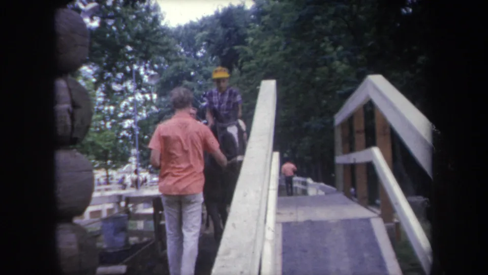 1968 Michigan frame showing a man beside a horse and rider in a yellow hat at a wooden ramp near a log structure.