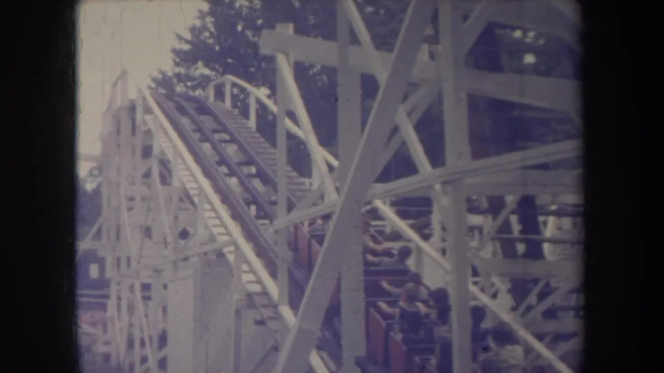 1968 Michigan home-movie frame with an ascending wooden coaster track, white supports and visible riders.