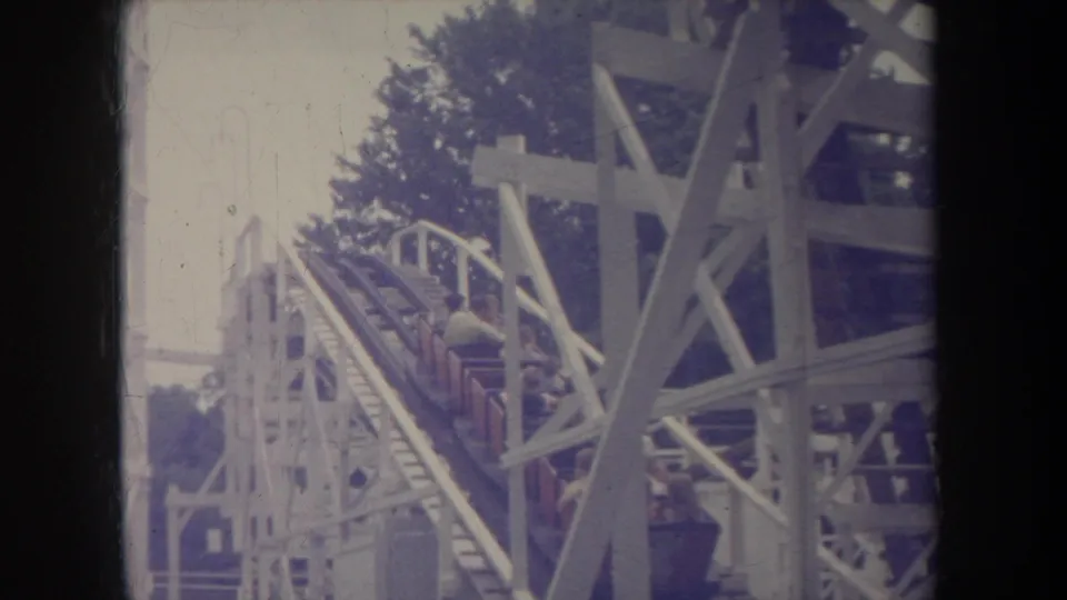 1968 Michigan home-movie frame of riders ascending a wooden roller coaster in red cars beside white supports.