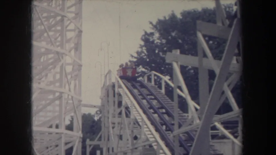 1968 Michigan 8mm view of a red roller coaster car moving up the lift hill through white wooden framework.