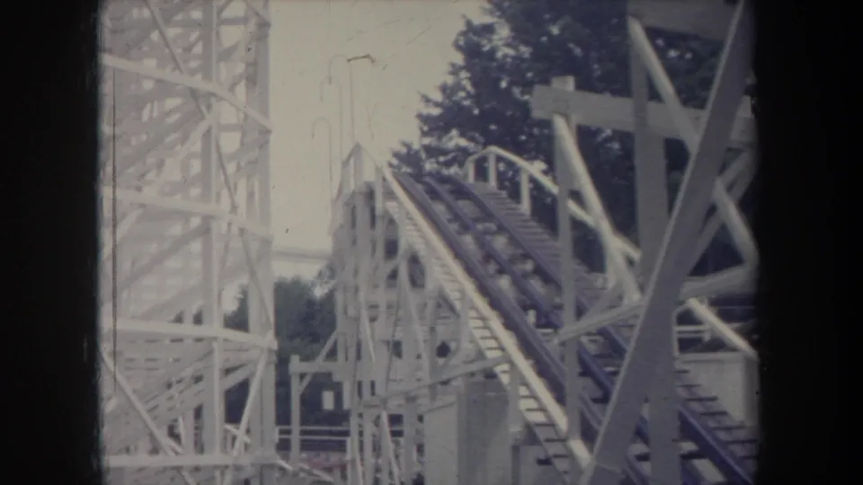 1968 Michigan home-movie frame showing the wooden coaster lift hill and surrounding white support structure after the car passes.