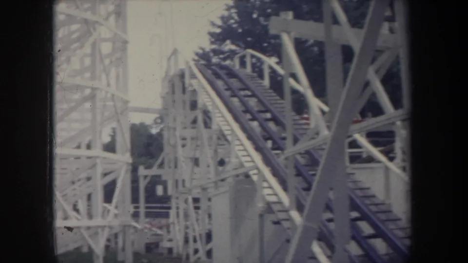 1968 Michigan home-movie frame showing a white wooden roller coaster lift hill, dark rails, support beams, and trees in the background.