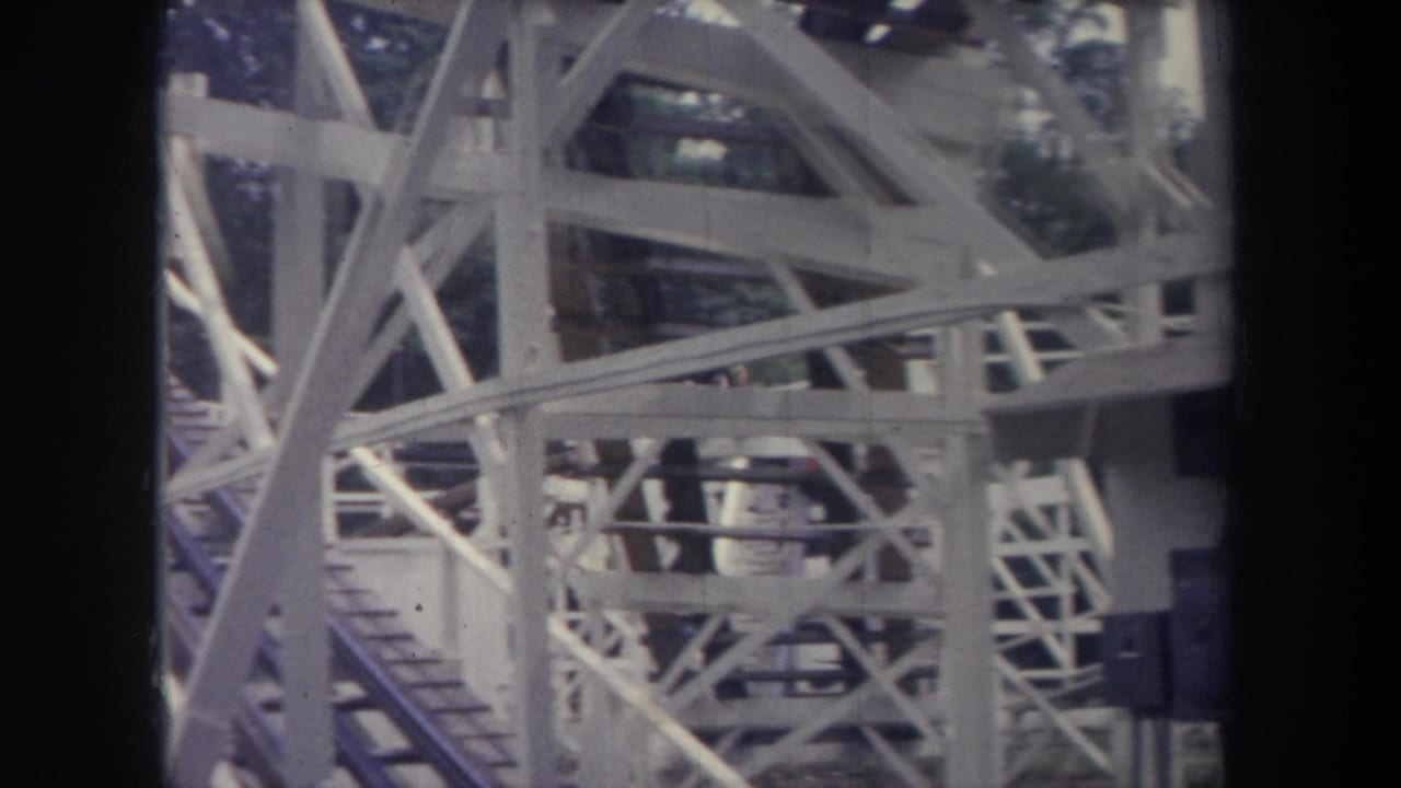 1968 Michigan amusement park frame with white wooden coaster supports and a red train carrying riders partly obscured by beams.